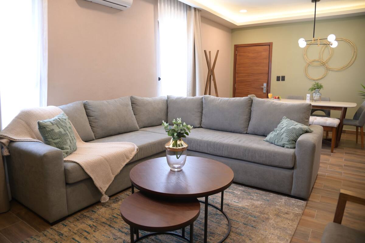 Een moderne woonkamer met een grijze bank, twee ronde salontafels, een houten deur en een eettafel met stoelen.