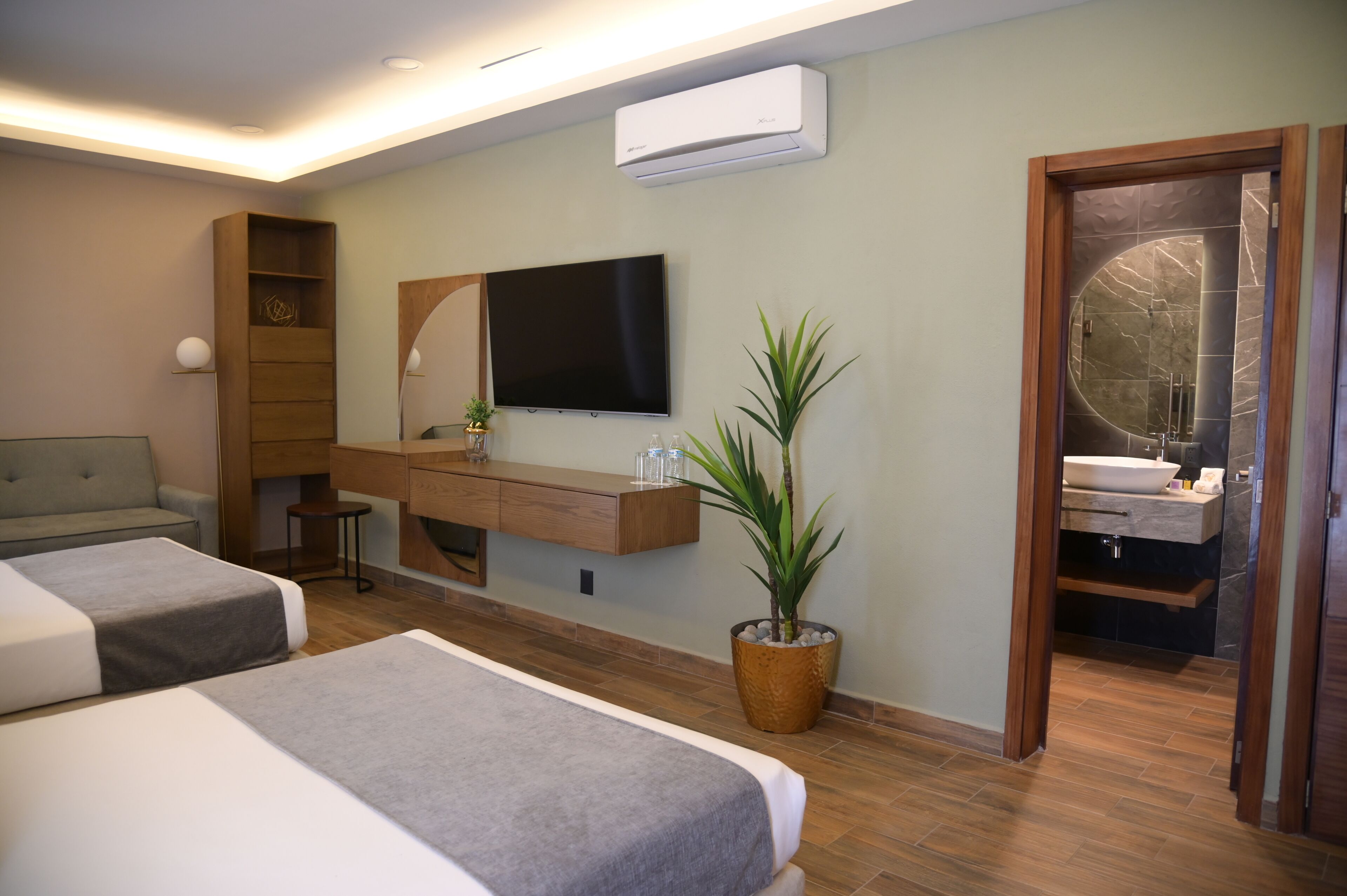 A hotel room with two beds, a TV, a wooden console table, a potted plant, and a bathroom visible through an open door.