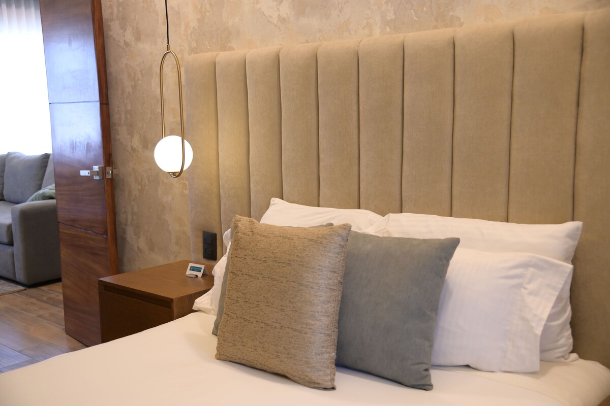A hotel room with a large bed, a wooden nightstand, a wall-mounted light, and a door to another room.