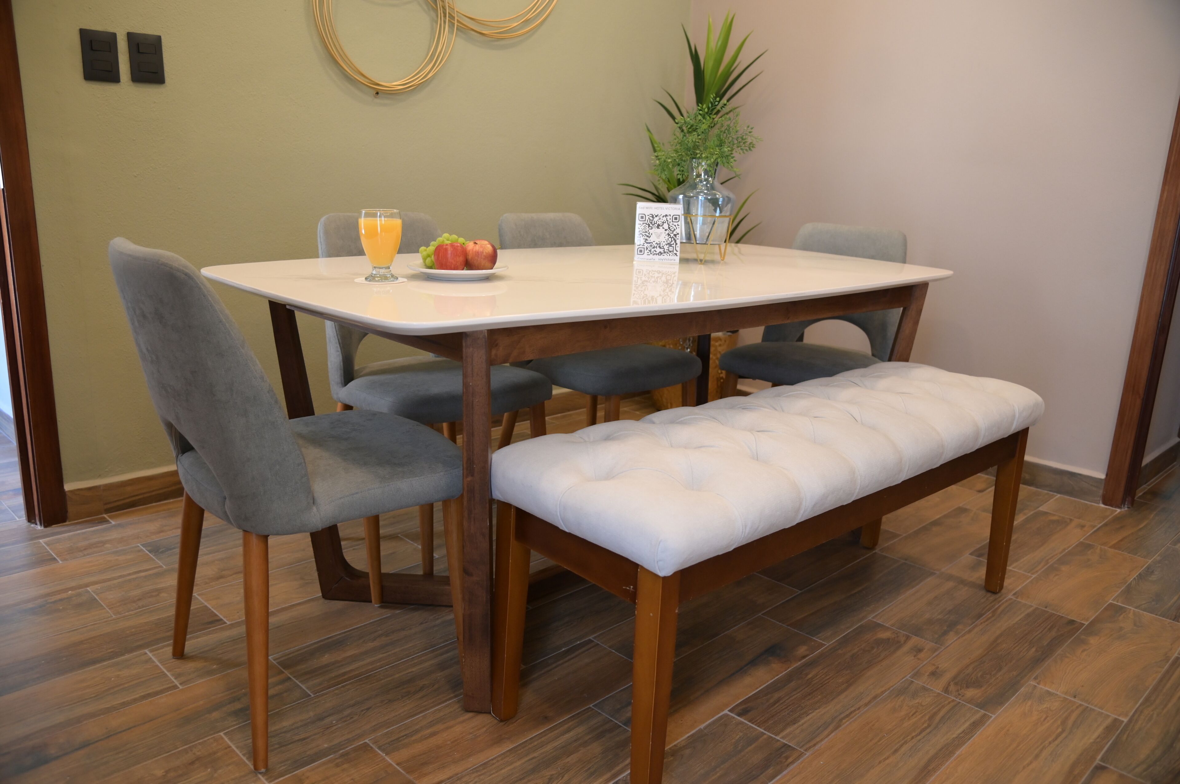 Un comedor con una mesa blanca, sillas tapizadas de color gris, un banco de madera y un jarrón con una planta.
