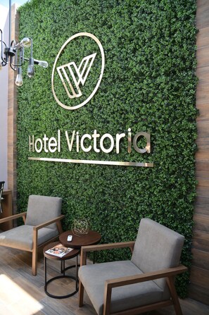 A modern hotel lobby with a green wall, a logo, and the name 'Hotel Victoria'. There are two grey armchairs and a small round table with a magazine and a decorative item. - Hotel Victoria (Lagos de Moreno)