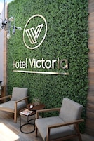 A modern hotel lobby with a green wall, a logo, and the name 'Hotel Victoria'. There are two grey armchairs and a small round table with a magazine and a decorative item.