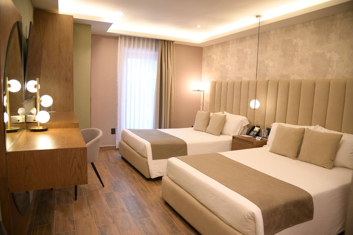 Een hotelkamer met twee bedden, een bureau met spiegel, een stoel, een nachtkastje met lamp en een raam met gordijnen.