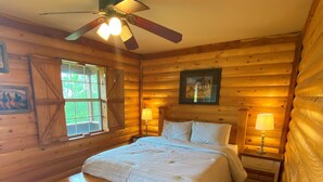 Camera da letto in una baita di legno, con un letto, due comodini con lampade, una ventola a soffitto e una finestra.