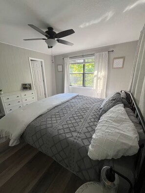 Un dormitorio con cama, ventilador de techo, ventana con cortinas, cómoda y radiador.