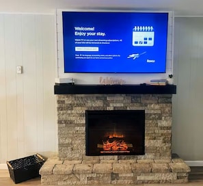 A flat-screen TV on a fireplace mantel with a welcome screen, a stone fireplace, and a small plant on the mantel. - Creekside Firefly Cottage (Bryson City)