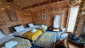 A cozy room with wooden walls, three beds with patterned bedding, a small table with cups, and string lights. - Pilgrims of the Goddess - Cabin (Glastonbury)
