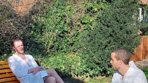 Two people sitting by a fire pit in a garden with a wooden bench and dense foliage in the background.