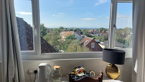A room with a view of a residential area through large windows, a table with a lamp, a chair, and a small table with a kettle and a glass pitcher. - Pilgrims of the Goddess - Room 8 (Glastonbury)