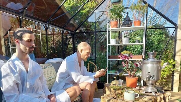 Two people sitting on a couch in a greenhouse with a wooden table and potted plants. - Pilgrims of the Goddess - Room 8 (Glastonbury)