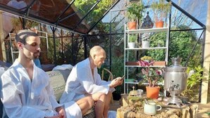 Two people sitting on a wooden deck under a glass structure with potted plants and a metal container. - Pilgrims of the Goddess - Room 7 (Glastonbury)