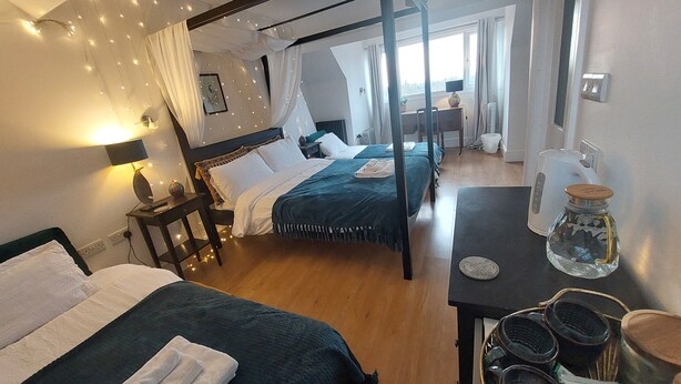 A bedroom with a four-poster bed, a bedside table with a lamp, and a view of the room through a window. - Pilgrims of the Goddess - Room 7 (Glastonbury)