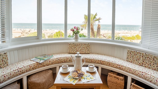 A bay window with a view of the beach, a bench with a patterned cushion, a table with a teapot, cups, and a plate of cookies.