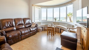 A living room with a leather sofa, a wooden coffee table, and a bay window with a view of the sea.