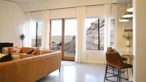 Een moderne woonkamer met een bank, een eethoek met stoelen, een keukenblad en grote ramen met gordijnen.