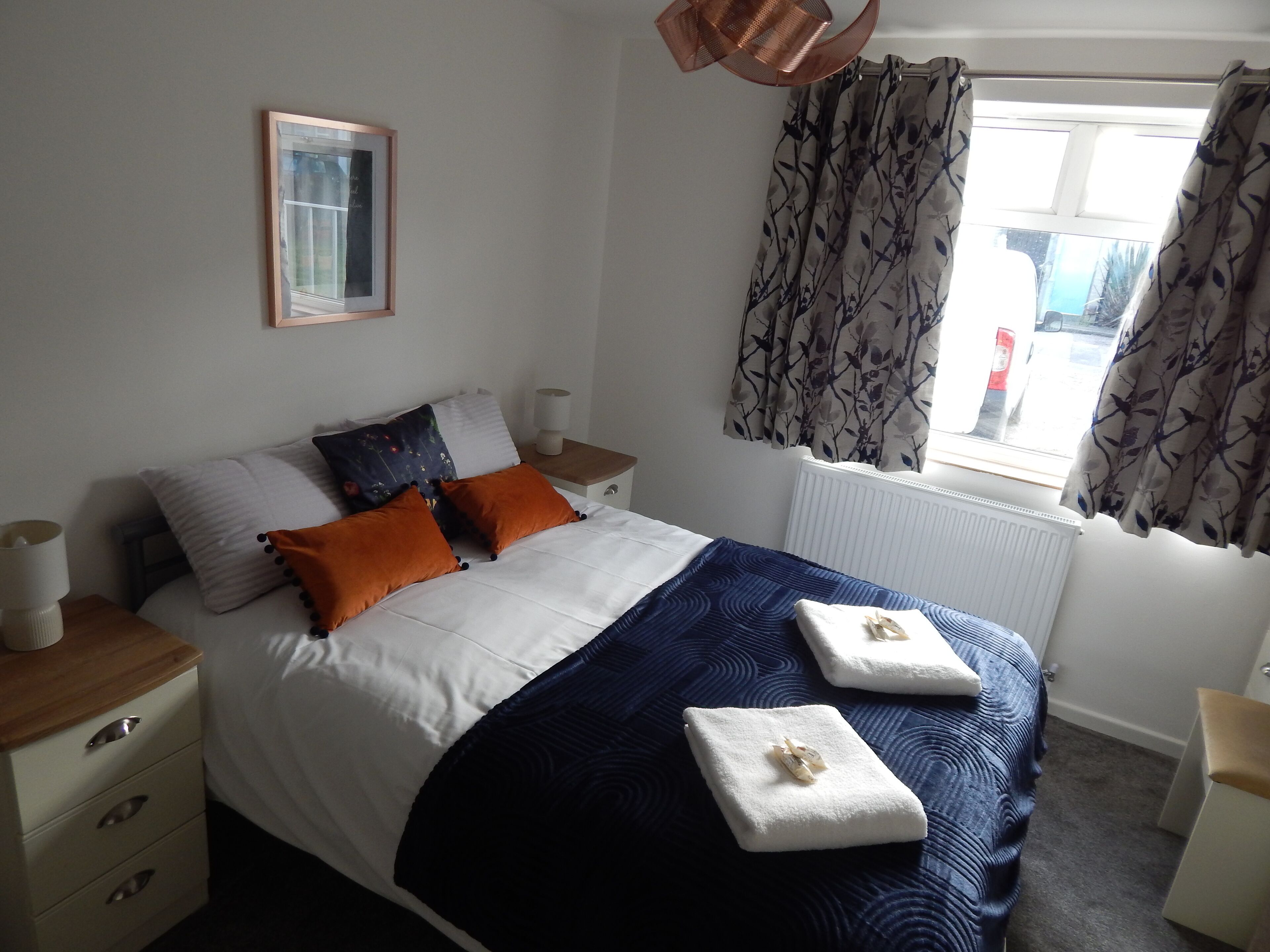 Een net opgemaakt bed met witte en marineblauwe beddengoed, twee oranje kussens en twee gevouwen handdoeken.