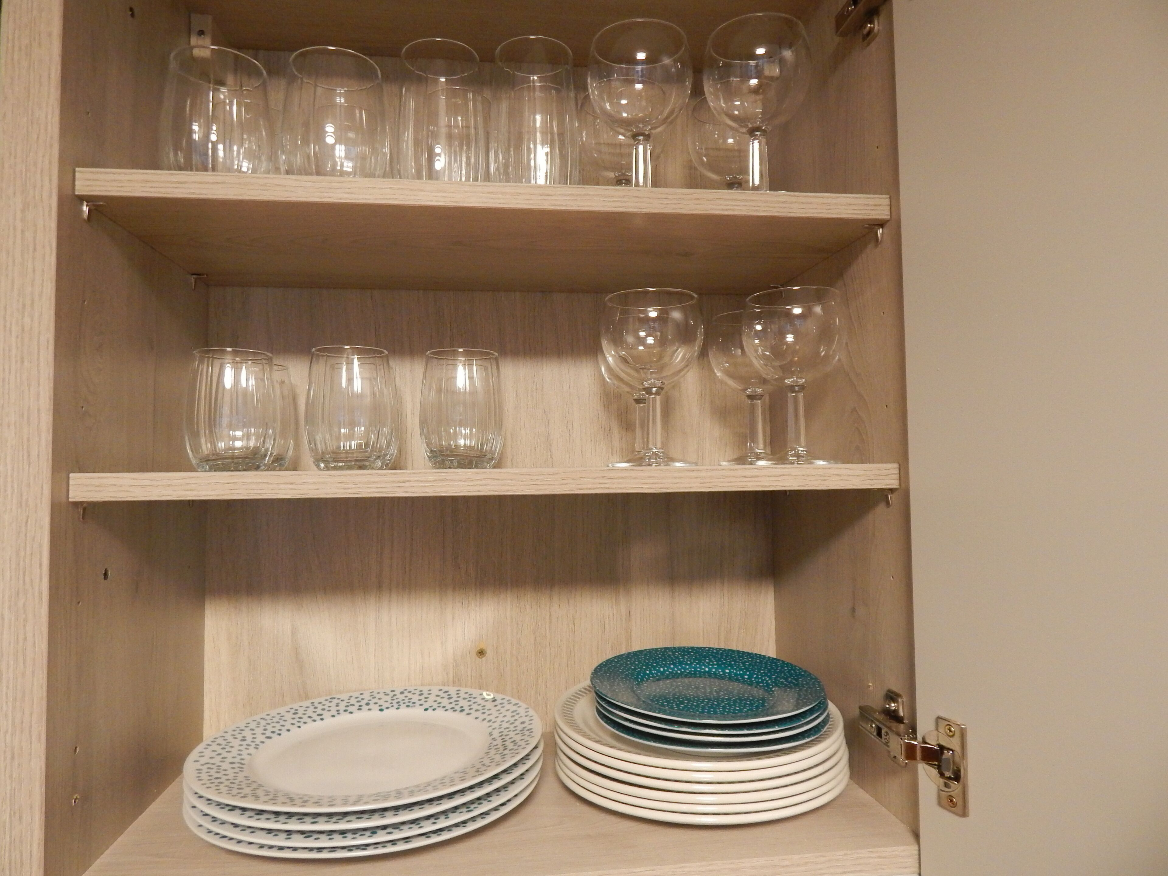 A wooden cabinet with shelves holding clear wine glasses and plates.