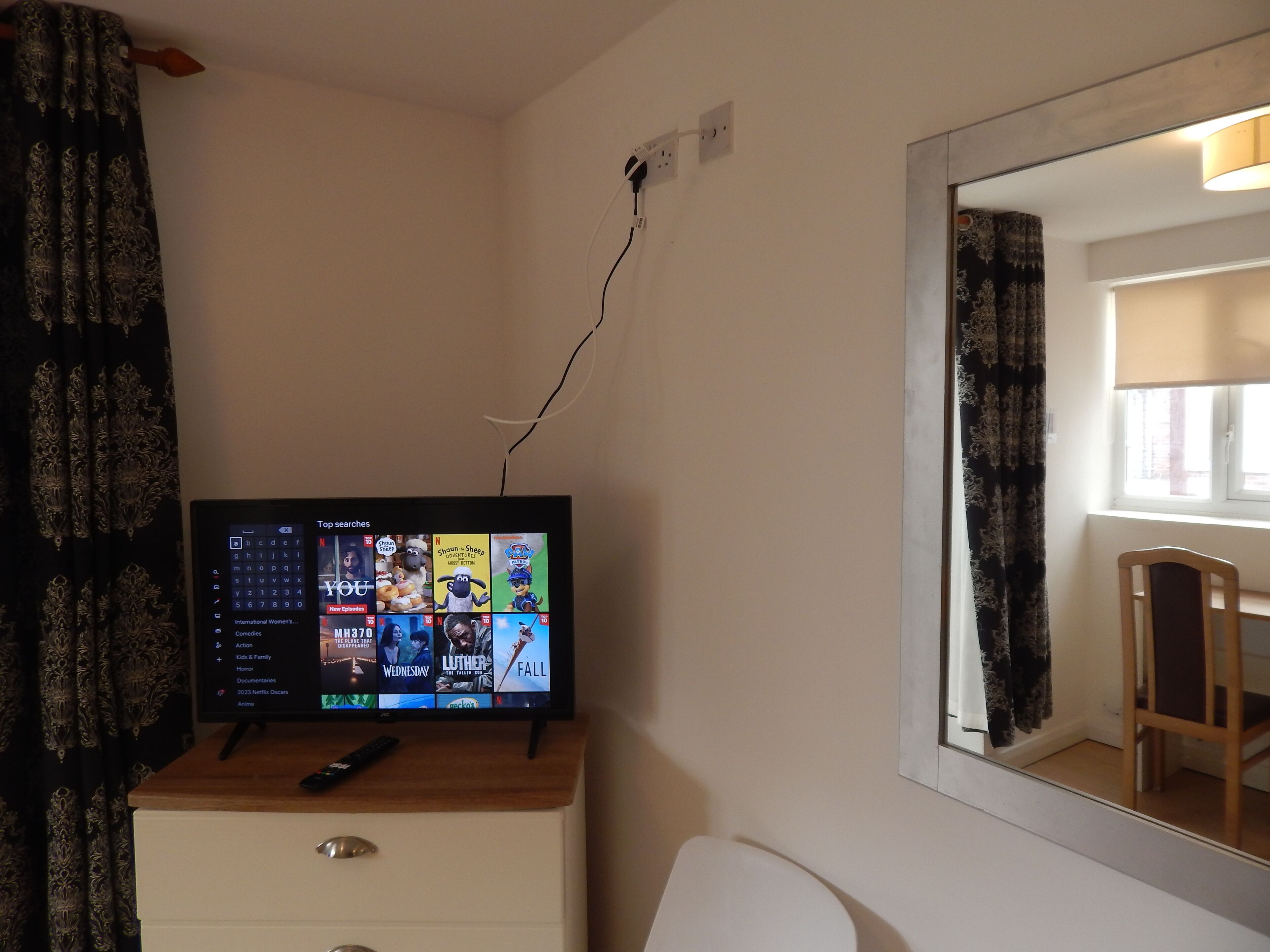 Une télévision posée sur une commode en bois, une télécommande à proximité, un miroir accroché au mur et une fenêtre avec des rideaux.