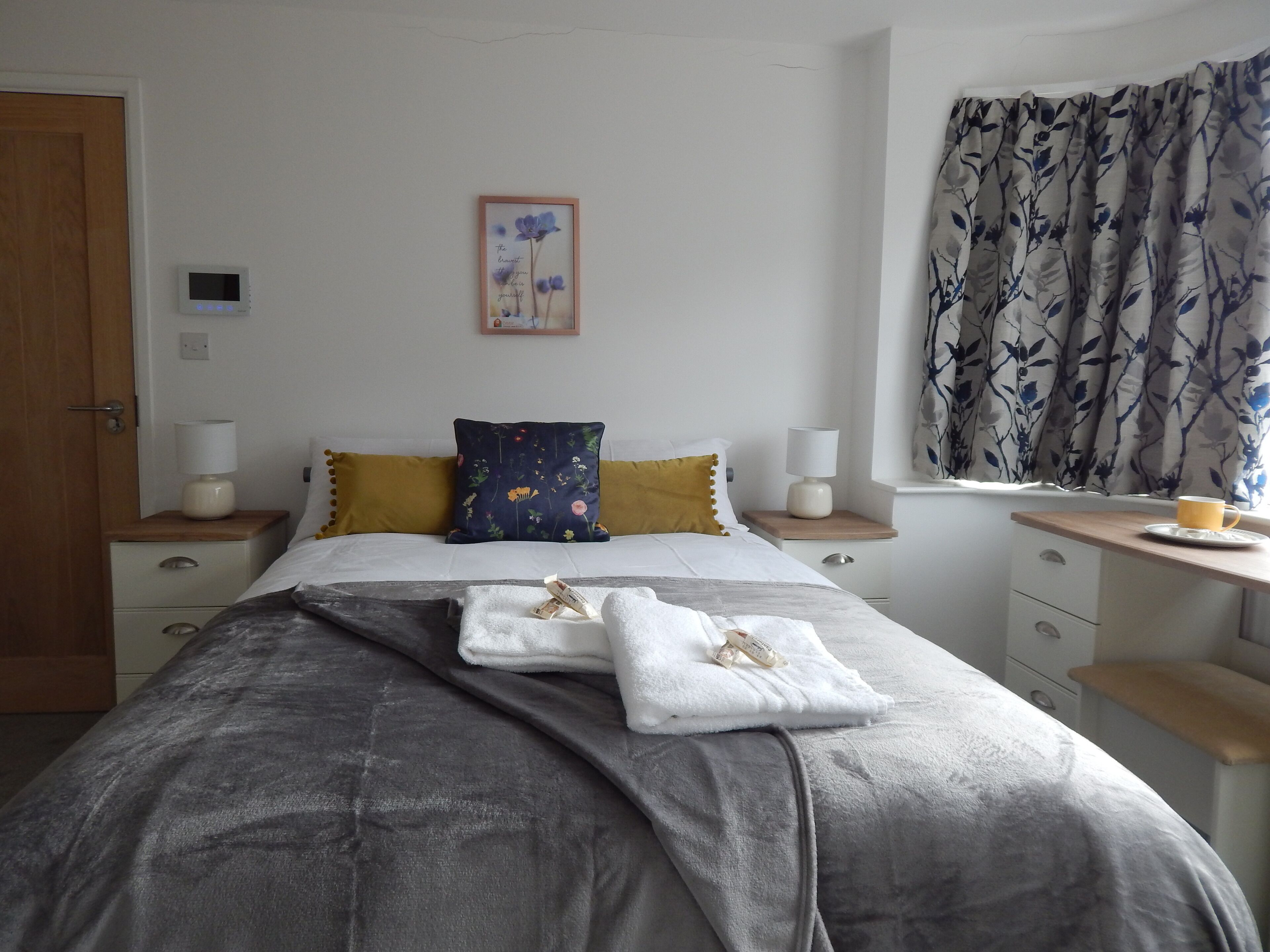 A neatly arranged bedroom with a bed, bedside tables, a nightstand, a window with curtains, and a wall-mounted thermostat.