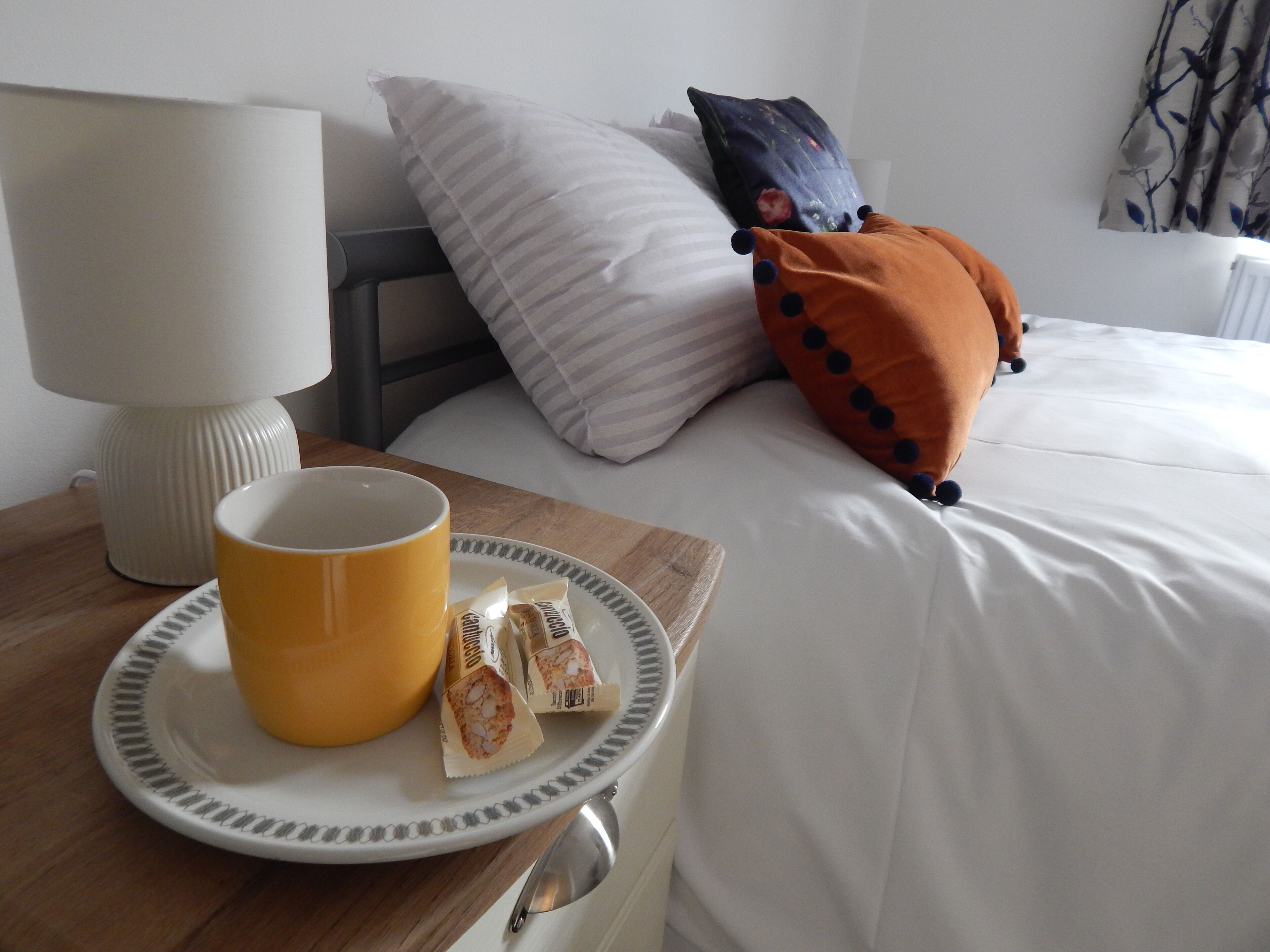 A neatly made bed with white bedding, a bedside lamp, a plate with a mug and snacks, and decorative pillows.