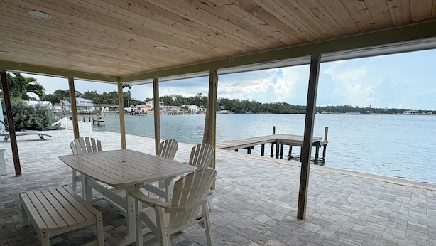 Une terrasse extérieure couverte, équipée d’une table et de chaises, une jetée s’avançant dans l’eau et une vue sur une côte lointaine.
