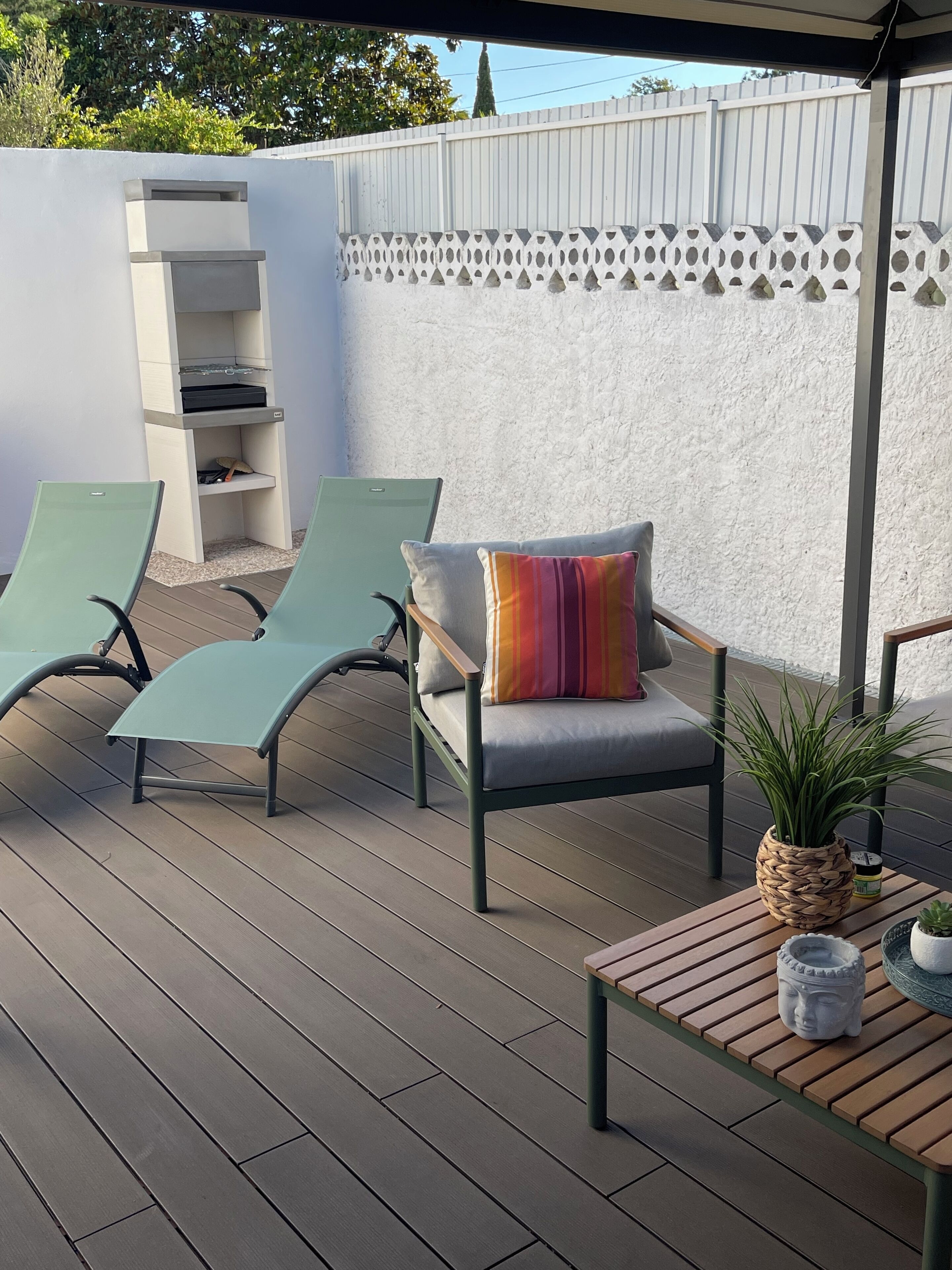 Un área de estar al aire libre cubierta con dos sillones, una butaca con cojín y una almohada colorida, una mesita con planta en maceta y una pared decorativa con diseño geométrico.