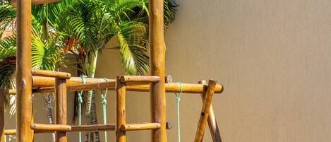 A wooden play structure with a slide and swings, set against a beige wall and surrounded by greenery.