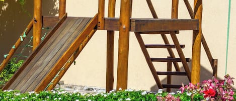 Eine hölzerne Spielkonstruktion mit Rutsche und Kletterelementen, vor einer Ziegelwand und umgeben von Grünpflanzen.