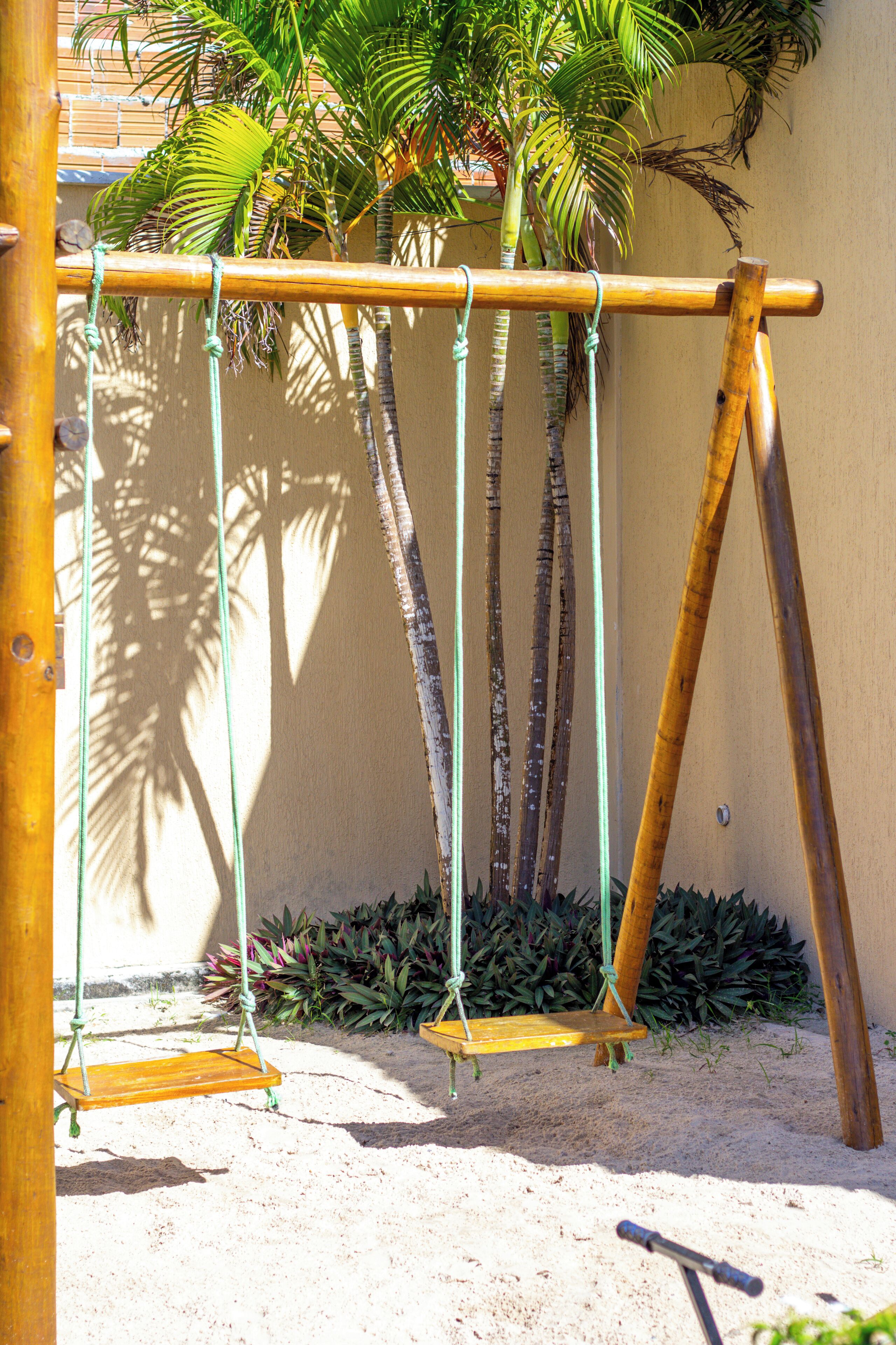 A wooden swing set with ropes, a sandy ground, and a palm tree in the background.