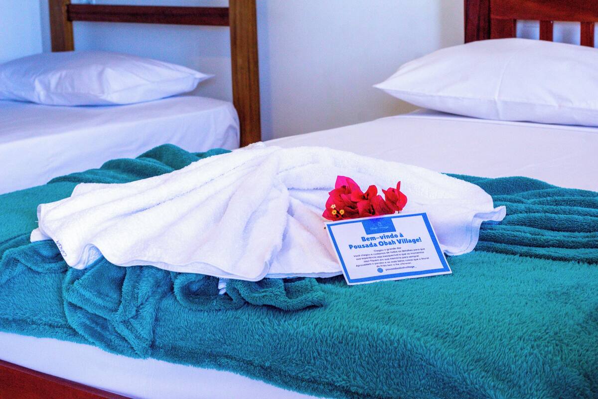 A hotel room with two beds, each with white linens and pillows. A teal towel is draped over one bed, and a white towel is on the other. A card with text is placed on the bed with the teal towel.