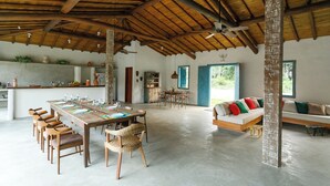 Un espacio amplio con techo de madera alto, una zona de comedor con una mesa larga y sillas, un sofá con cojines de colores, y vistas al exterior a través de una ventana.