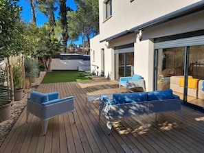 A modern outdoor patio with wooden flooring, two blue cushioned chairs, and a glass sliding door leading to a living area.