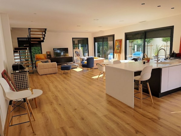 A modern open-plan living area with a kitchen, wooden flooring, and large windows.
