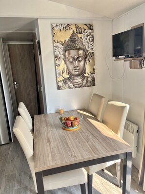 A dining area with a wooden table, white chairs, a fruit bowl, and a Buddha painting on the wall. - Mobile home Zen 40 m2 in Frejus in the Var for up to 8 people (Fréjus)