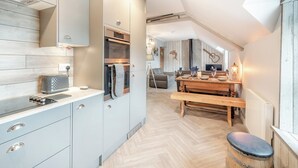 Ett modernt kök med en inbyggd ugn, en matplats med ett träbord och en träbänk samt ett sovloft med ett sluttande tak.