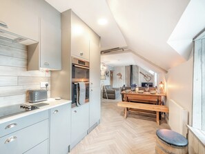 Una cuina moderna amb forn integrat, zona de menjador amb taula i banc de fusta, i un dormitori amb àtic i sostre inclinat.