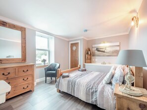 Un dormitorio con un aparador de madera, una silla, una cama con manta a rayas, un estante montado en la pared con libros, un cuadro y una lámpara.
