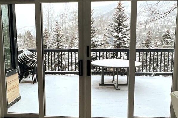 A snow-covered deck with a table and chairs, a wooden deck area, and a view of snow-covered trees and mountains.