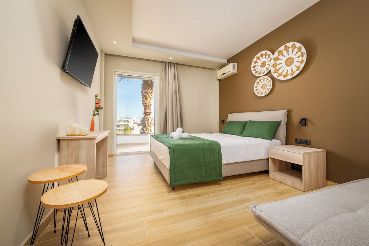 A modern hotel room with a bed, bedside tables, a TV, a balcony with a view, and a dining area with stools.