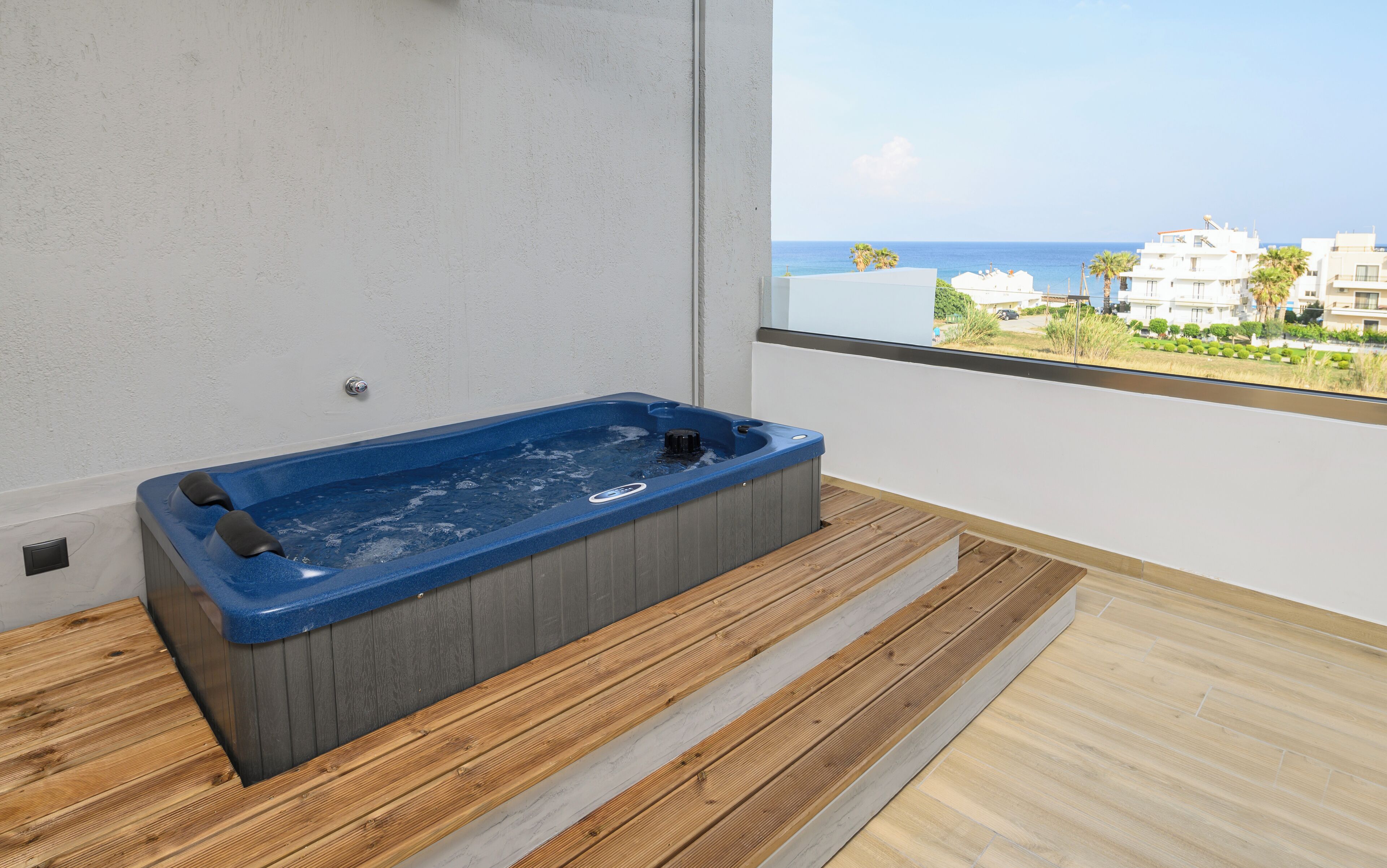 Un baño moderno con suelo de madera, una bañera de hidromasaje azul y vistas al mar y edificios.