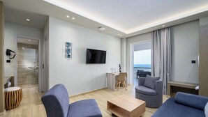 A modern living room with a flat-screen TV, grey sofas, a wooden coffee table, and a view of the sea through large windows. - Nicolas Grand Suites - Adults Only (Kos)