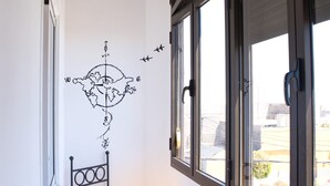 Una habitación en esquina con ventana, un adorno de brújula en la pared, una silla amarilla y una mesita con una copa de vino y una revista.