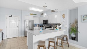 A modern kitchen with white cabinetry, stainless steel appliances, a central island with stools, and a decorative plant. - Gulf Crest Gulf-Front Condo | Beach Chairs Included | Sleeps 8 (Panama City Beach)