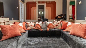 A living room with a dark grey sectional sofa adorned with patterned and solid orange pillows, a black armchair, and a small table with a lamp. A doorway leads to a bathroom with a red shower curtain.