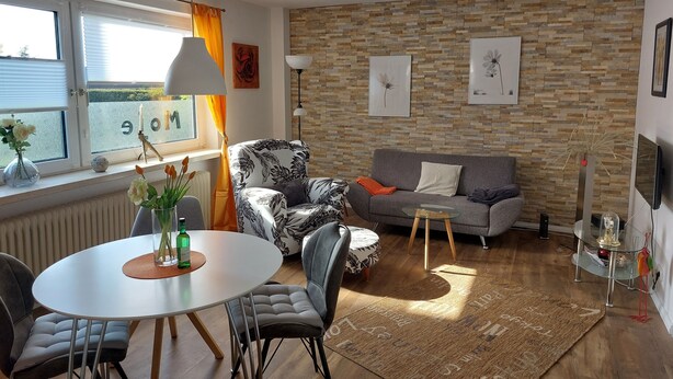 Uma sala de estar moderna com parede de pedra, sofá cinzento, mesa de jantar redonda e uma janela grande com cortinas.