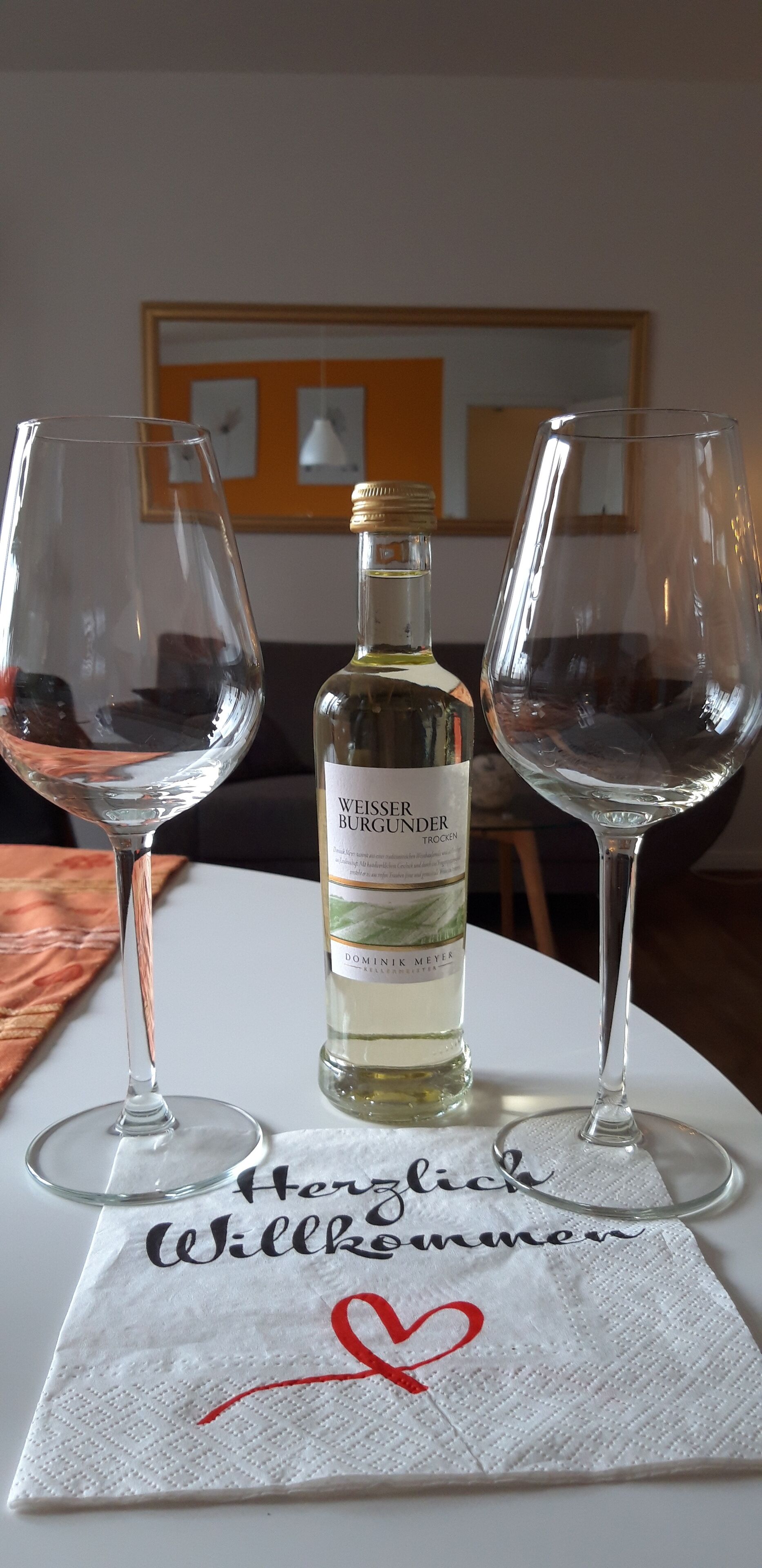 Two empty wine glasses, a bottle of Weiss Burghunder Trocken, and a napkin with "Herzlich Willkommen" and a red heart.