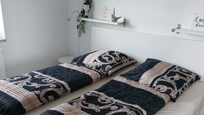 Quarto com duas camas, cada uma com roupa de cama estampada e um travesseiro decorativo. Há decorações nas paredes, uma estante com objetos e uma janela.