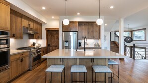Una cocina moderna con armarios de madera, electrodomésticos de acero inoxidable, una isla central con taburetes altos y luminarias colgantes.