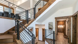 Una escalera de madera de varios niveles con barandillas de metal, que conduce a una zona de cocina con armarios de madera y suelo de baldosas.