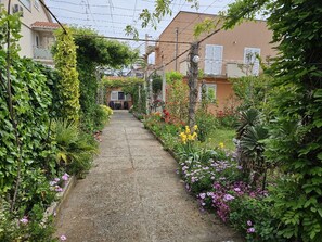 Un étroit sentier bordé d’une végétation luxuriante et de plantes en fleurs, qui mène à un bâtiment doté d’un balcon.